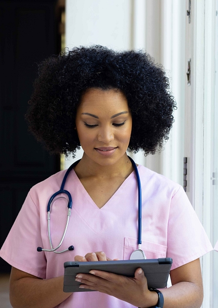 Young biracial female nurse wearing pink scrubs checking tablet at home WEB