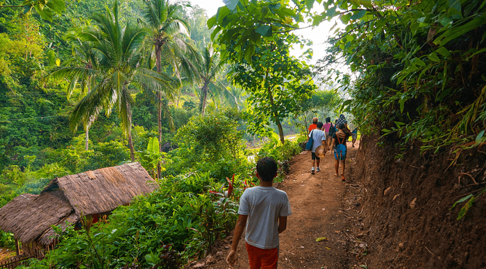 Group of hikers walking on tropical dirt trail past thatched hut surrounded by lush palm trees and vegetation